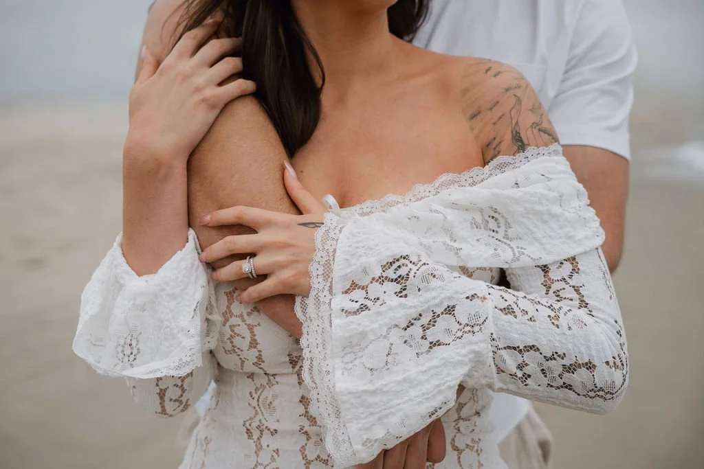 Close-up of a woman in a white lace off-shoulder dress, holding her shoulder with one hand, with tattooed arms and a wedding ring visible. The person's face is not visible, and there is a beach background.