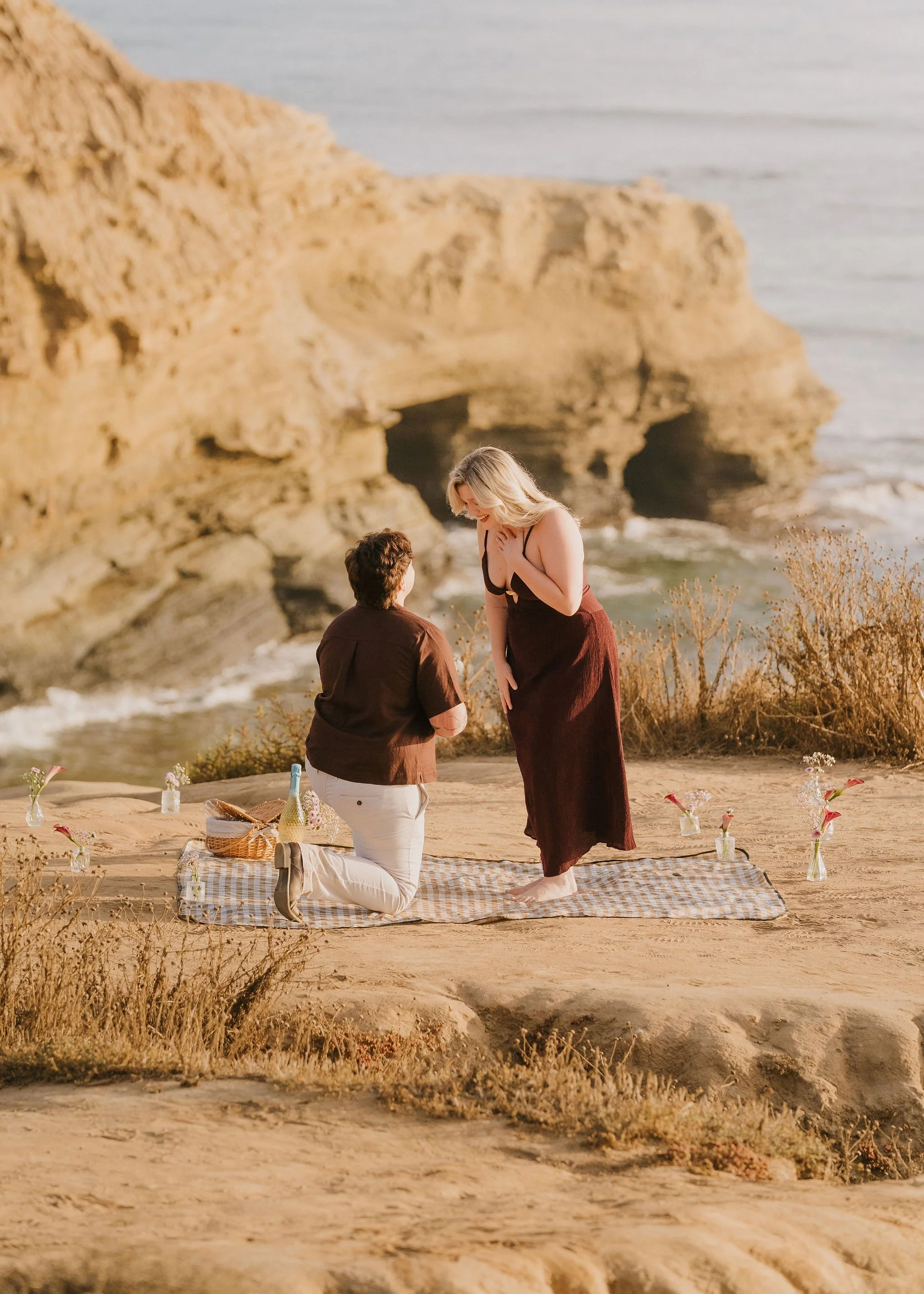 Ash kneeling to propose to Megan on a picnic blanket at Sunset Cliffs, San Diego, with the ocean and cliffs behind them