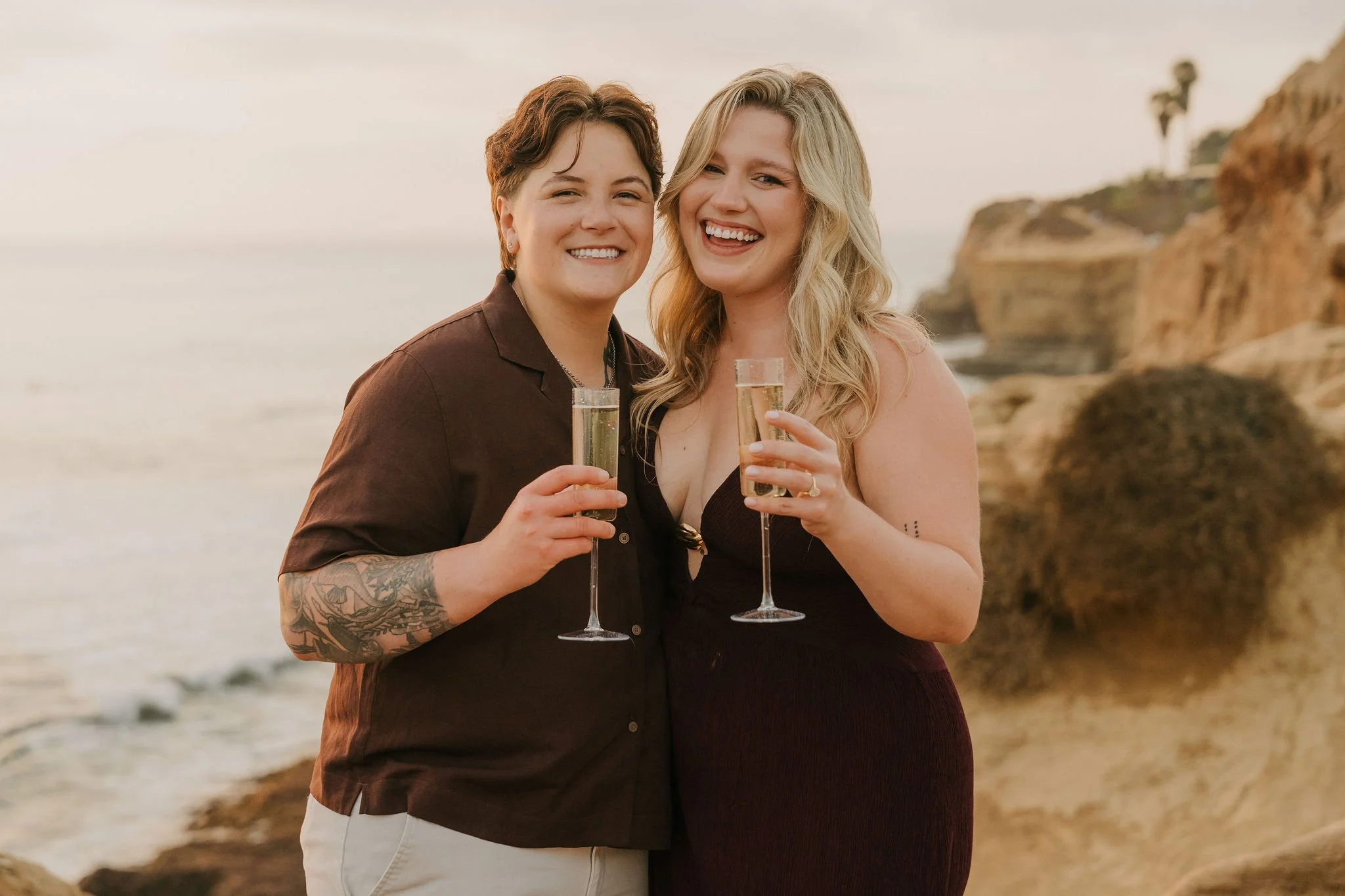 Queer couple toasting champagne after their surprise engagement at Sunset Cliffs, San Diego, with the ocean and cliffs behind them