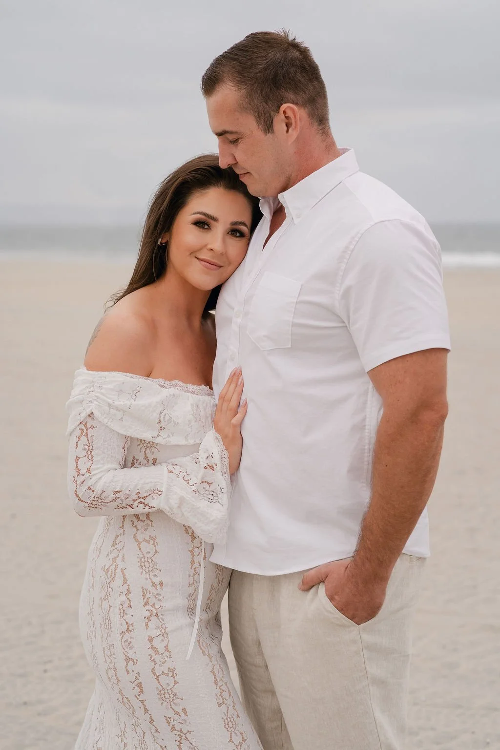 A man and woman standing close together on a beach, smiling at the camera. The woman has long dark hair and is wearing an off-shoulder white lace dress. The man has short hair and is dressed in a white shirt and beige pants, with his hand in his pock