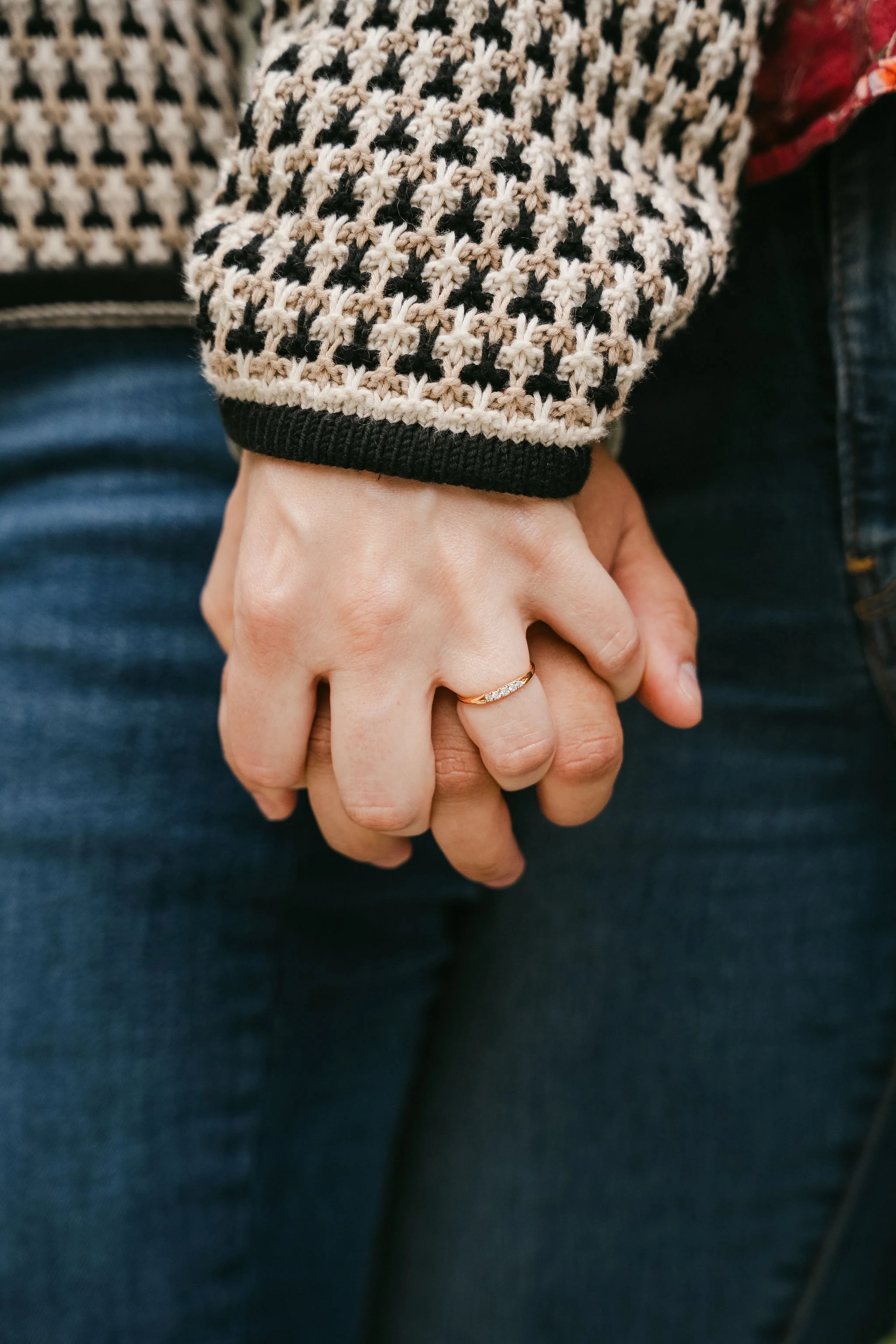 Close-up of a hand with a gold ring holding another hand, with a person wearing a black and white patterned sweater sleeve in the background.