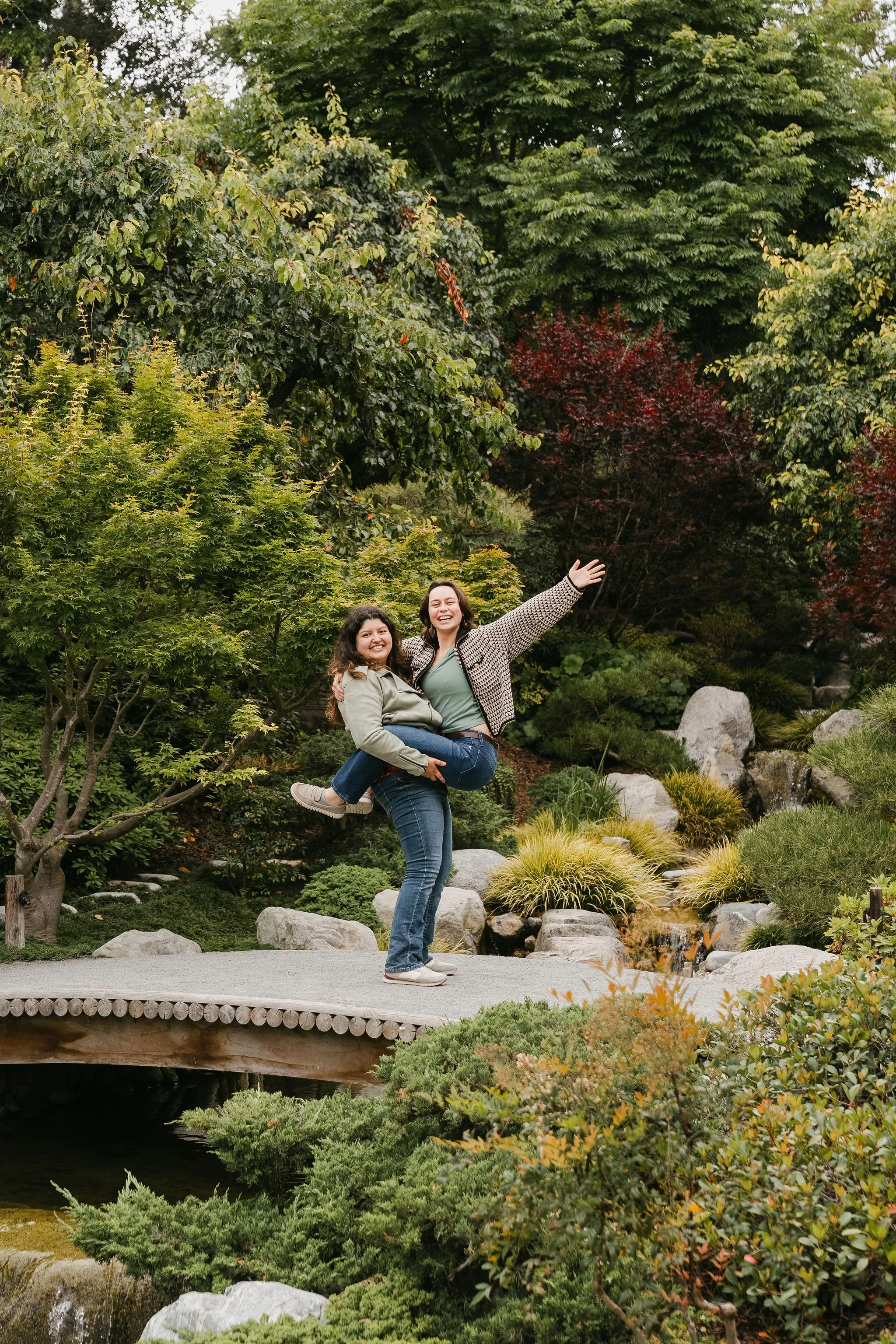 Two women smiling and posing in a lush, green garden with trees, shrubs, rocks, and a small waterfall.