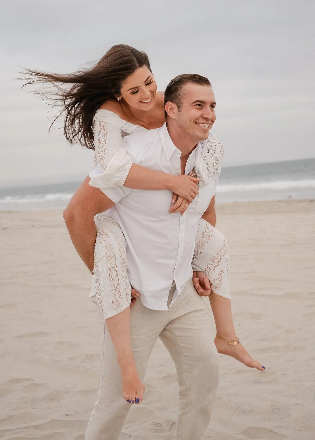 A couple enjoying time together at the beach, the woman on the man's back, both smiling and dressed in white. The ocean and cloudy sky are in the background.