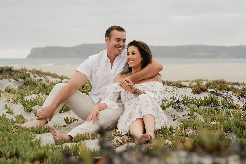 A smiling couple sitting on the beach among green succulent plants with an ocean and cloudy sky in the background.