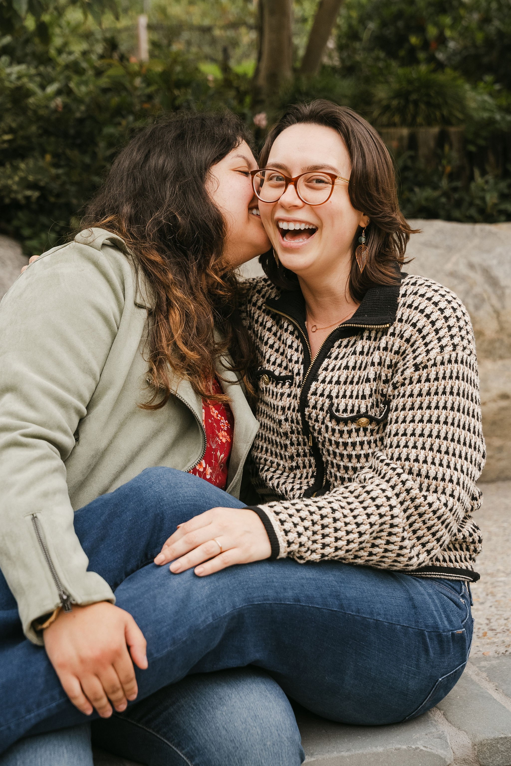 Two women sitting outdoors, one whispering to the other, both smiling and laughing in a park setting with greenery and rocks.