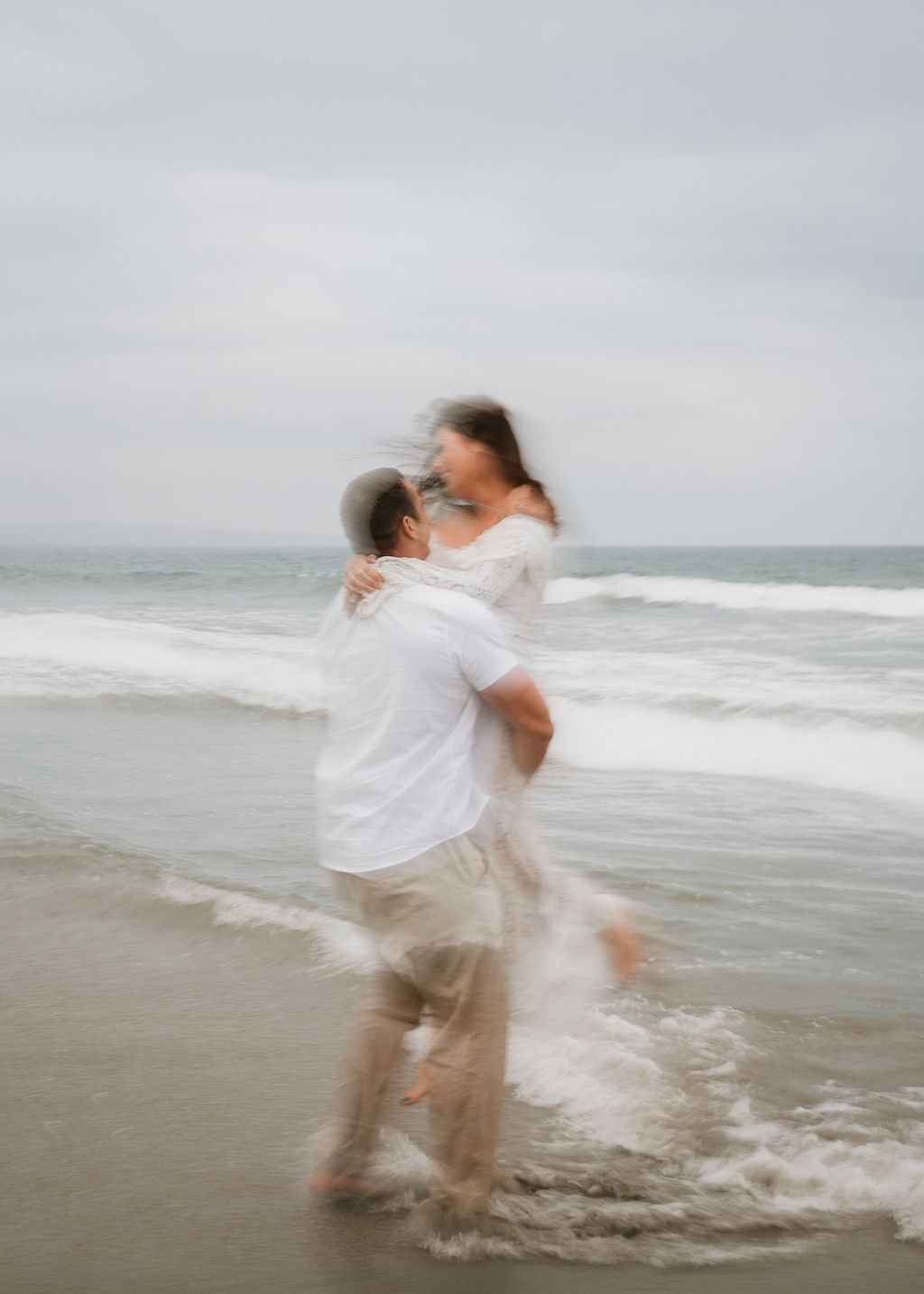 A couple at the beach during a cloudy day, with the man lifting the woman in his arms as they stand in the shallow water, embracing each other.