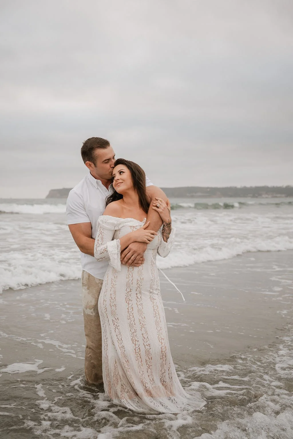 A couple embracing on the beach during cloudy weather, with the ocean and distant cliffs in the background.