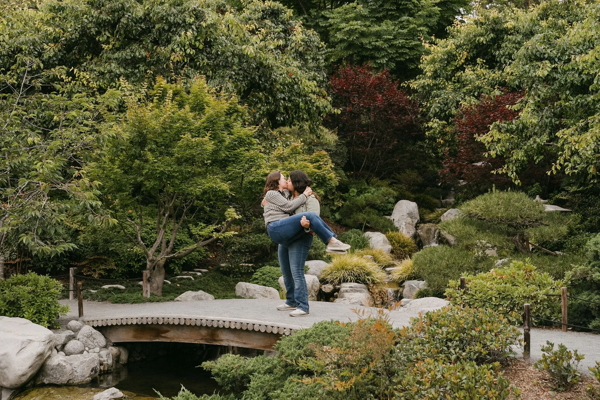 Two women sharing a kiss in a lush outdoor garden with green trees, bushes, rocks, and a small bridge over a pond.