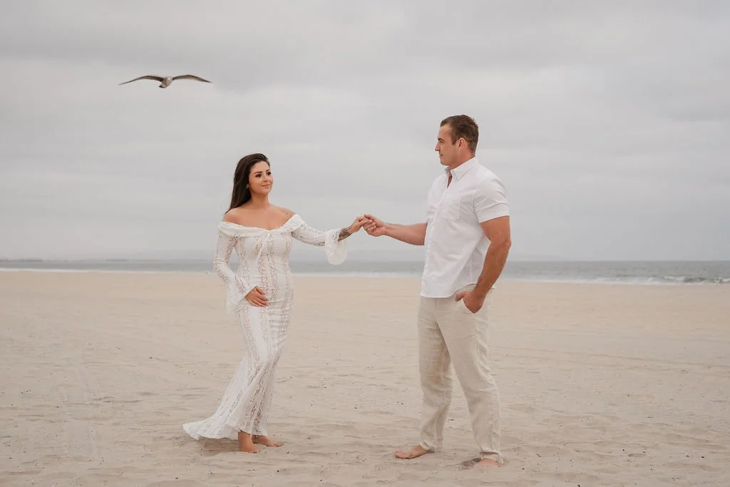 A woman in a white lace dress and a man in a white shirt and beige pants standing barefoot on a sandy beach, holding hands with a seagull flying in the overcast sky in the background.