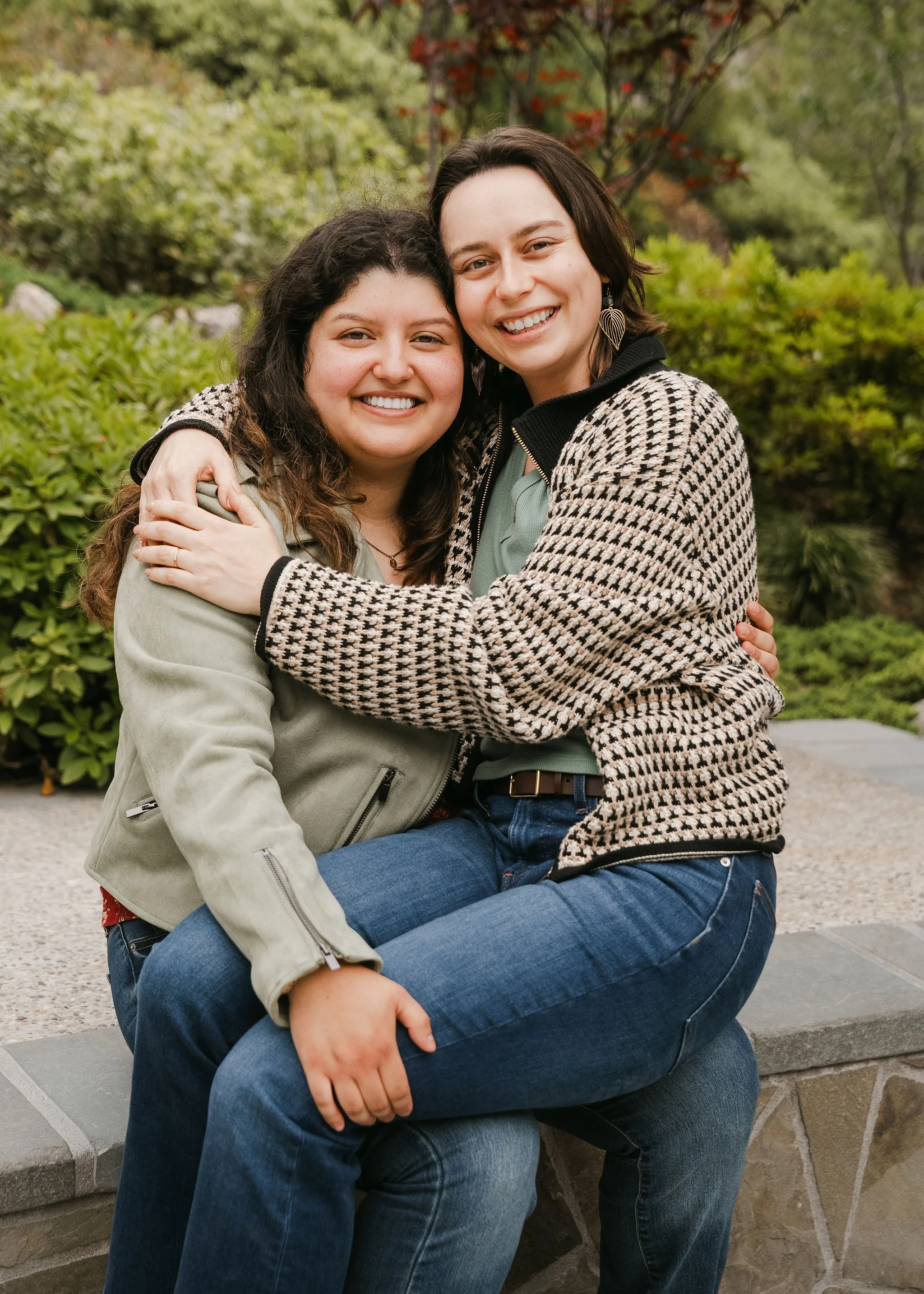 Two smiling women embracing each other outdoors, sitting on a stone bench surrounded by greenery with trees in the background.