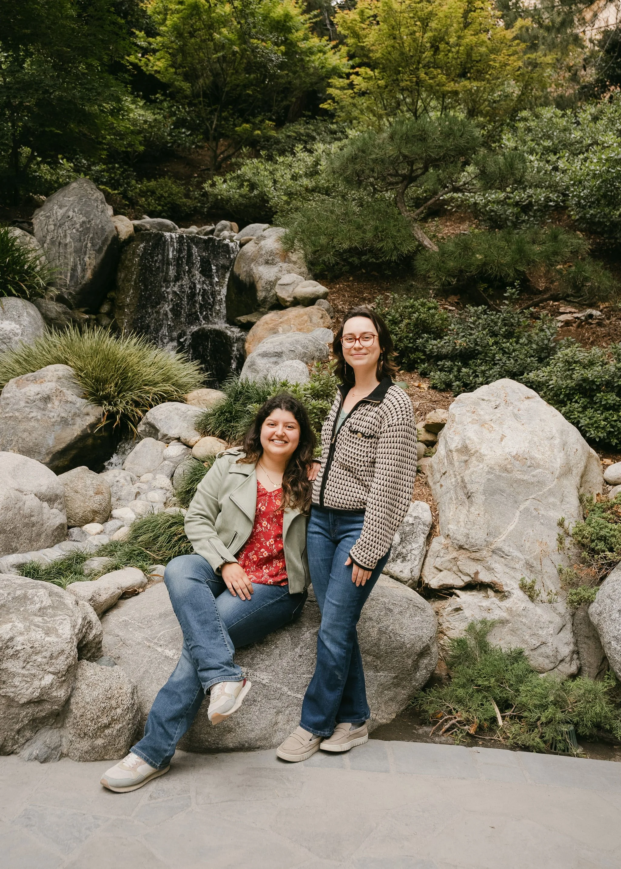 Two women smiling and posing in front of a small waterfall and rocks in a lush, green garden with trees and shrubs.