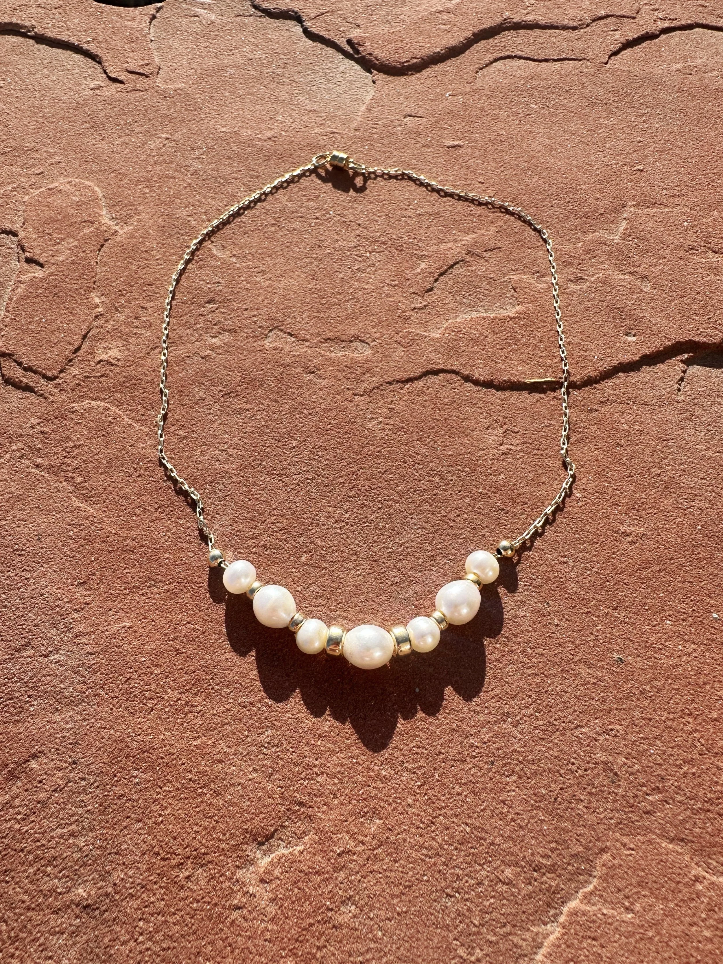 The Vesper Necklace flat lay on red stone — gold-filled baroque pearl necklace with paperclip chain and magnetic clasp in sunlight