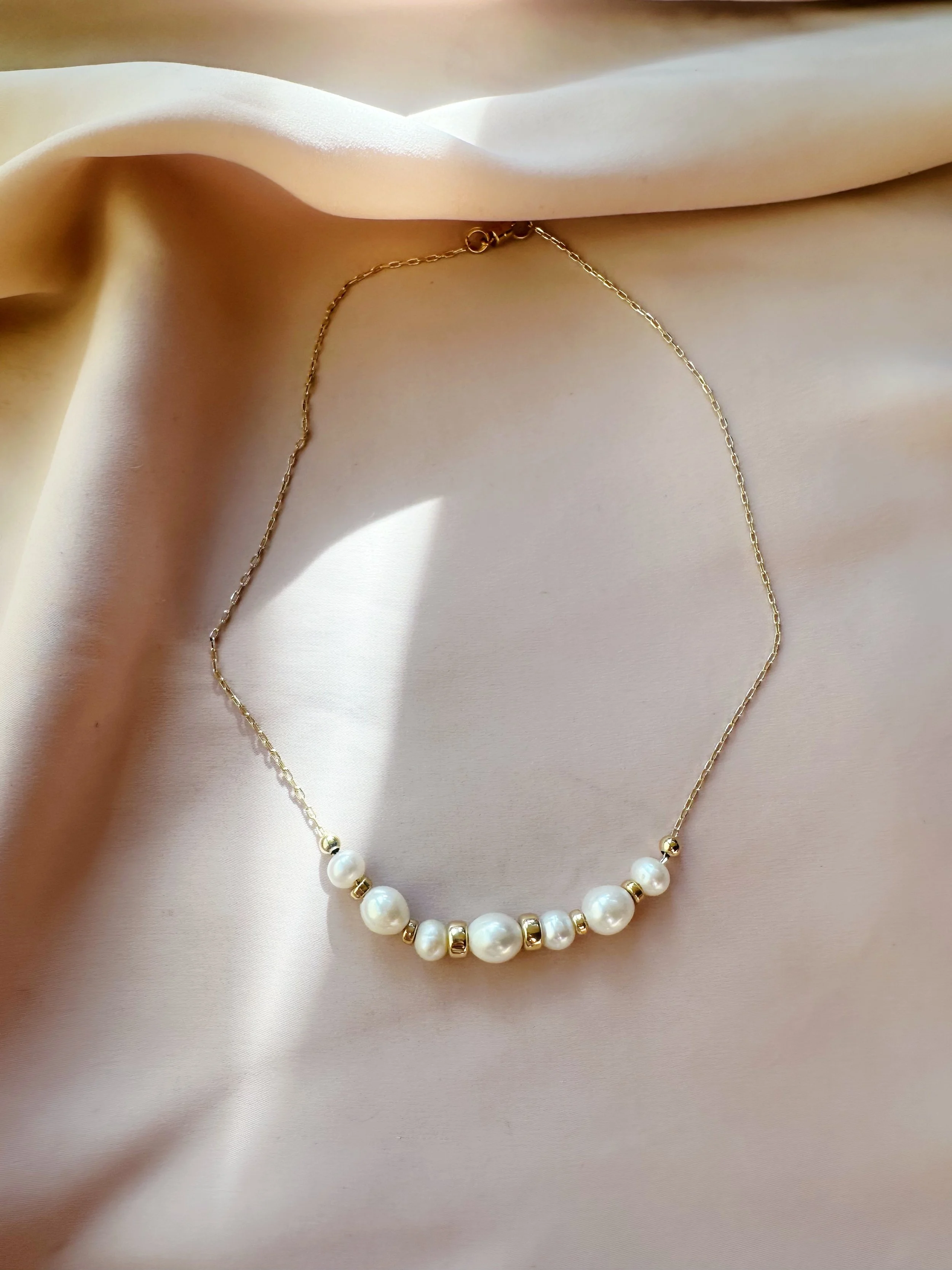 The Vesper gold-filled pearl necklace laid on cream silk — dainty paperclip chain with baroque pearl and gold bead detail, overhead flat lay