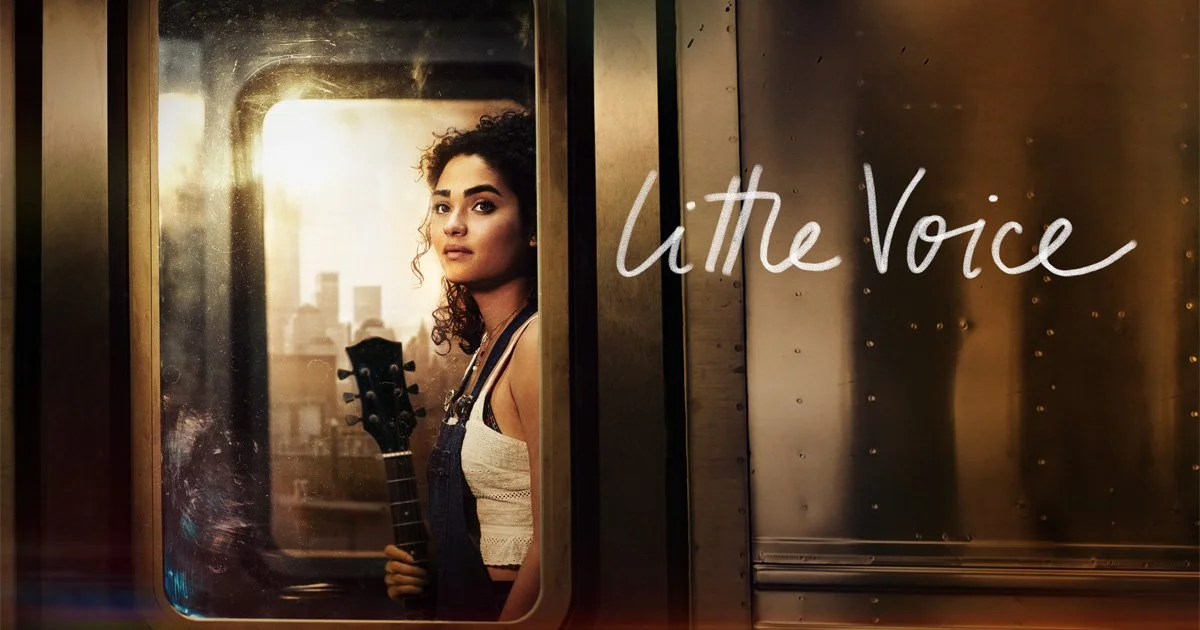 Wide angle shot of main character Bess on a subway train looking out of the window. She is mixed race and has light skin. Her hair is curly and tied up. She is holding a guitar. On the right, in white cursive the title reads ‘Little Voice’