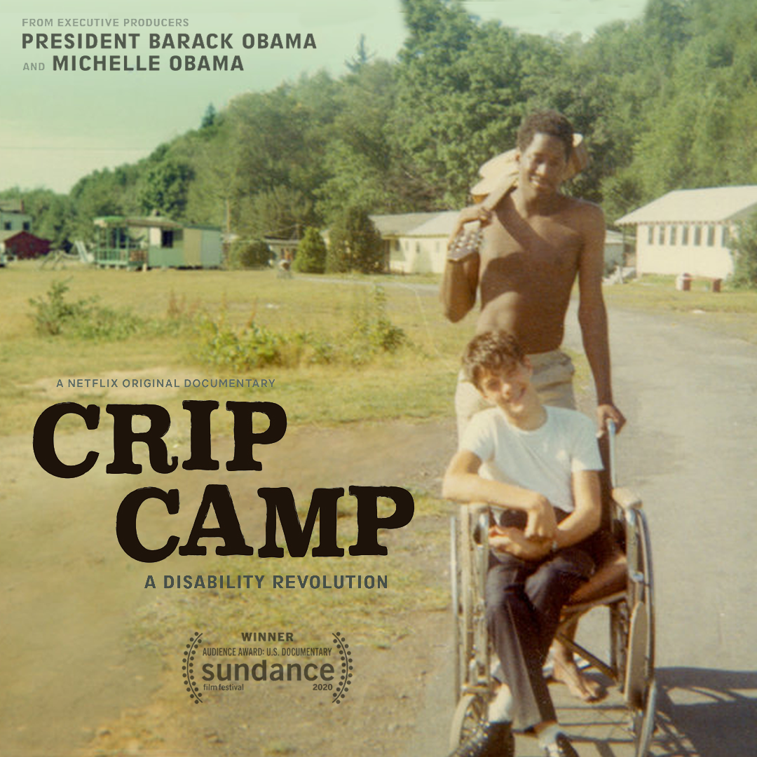 An African American counselor pushes a white camper in a wheelchair in with one hand, whilst holding a guitar with his shoulder over the other. Camp buildings fill the background. 