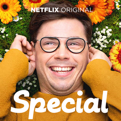 portrait of Ryan O’Connell - he is looking into the camera and smiling. He has dark brown hair, brown eyes, white skin and wears glasses. He is laid on grass and flowers, holding his hands up by his face.