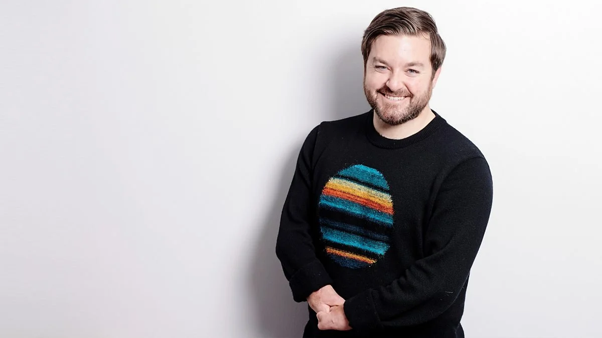 Alex is stood against a white background. He his clasping his hands together in front of his waist, smiling into the camera. He has white skin and light brown hair. He is wearing a black jumper with a colourful circle graphic.