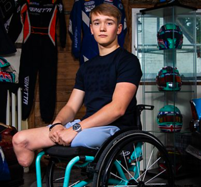 Portrait of Billy in his manual wheelchair. He is white, with blond hair. He is wearing a black t-shirt. His racing helmets and suits are in the background.