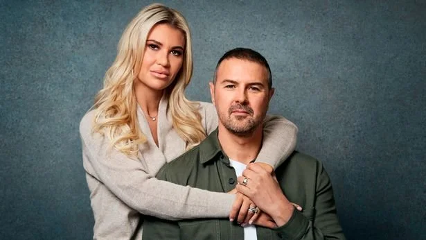 Against a grey background, on the left is Christine, She is white with blonde hair. She has her arms around Paddy on the right - he is white with short dark hair.