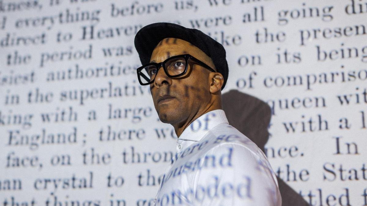 A picture of Jay Blades. He's a black man, wearing glasses and a black flatcap with a white shirt stood in front of a projector, with words displayed.