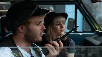 A close up through a car window of a white man (Alan) holding a walkie-talkie. In the background, a white woman with blonde hair is wearing a black hat, looking through the windscreen.