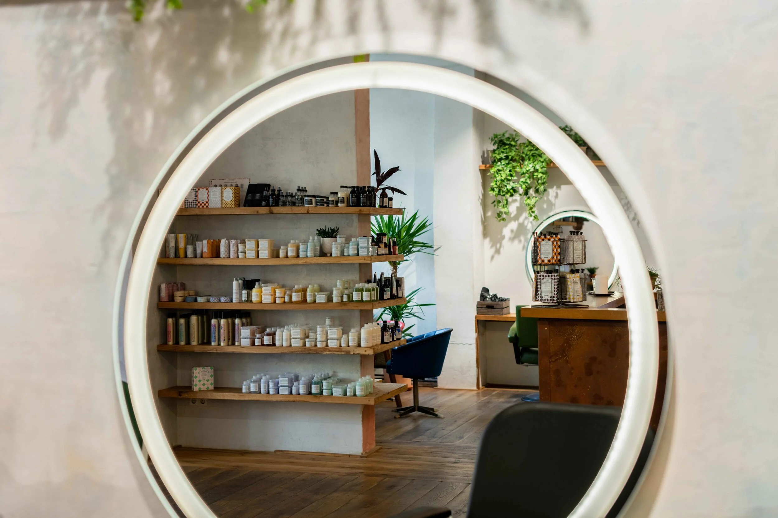 A hair salon interior reflected in a round mirror, showing shelves with various hair products, a desk with a chair, and plants.