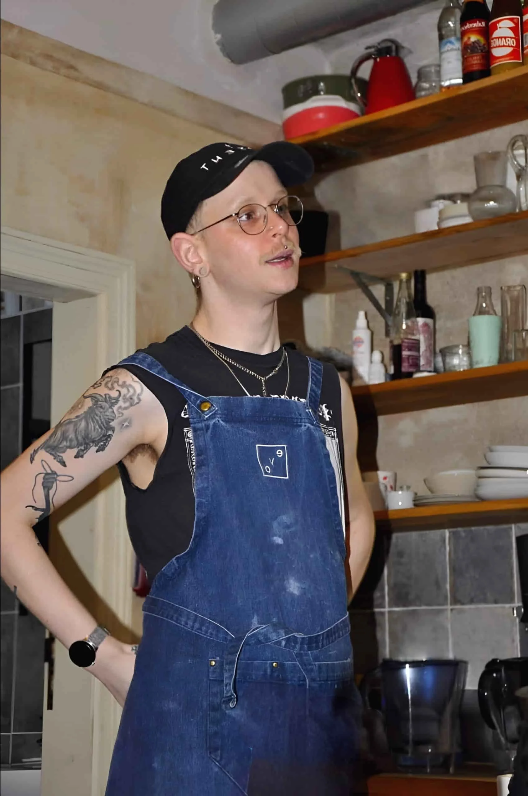 Tom wearing glasses and a black cap, standing in the salon backroom in a denim apron.