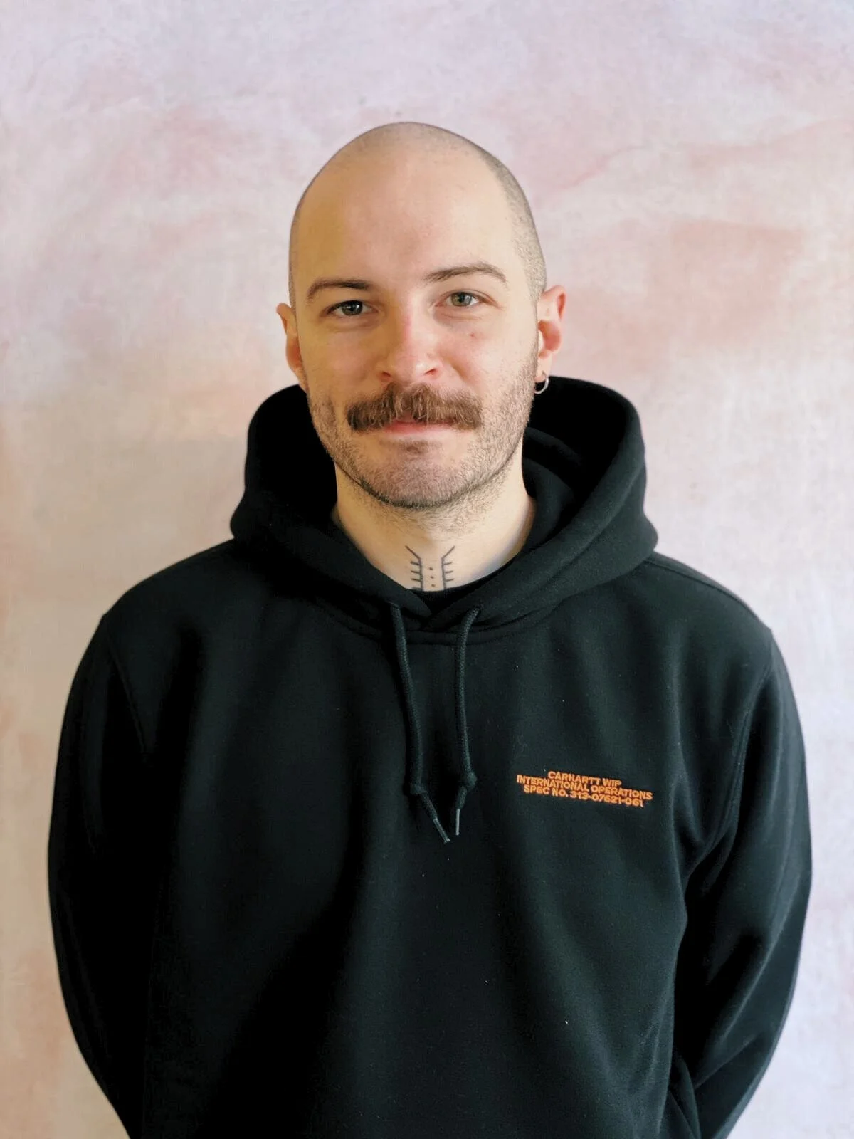 NeoBerlin salon manager Dale with a shaved head and mustache, standing in front of a light pink wall.