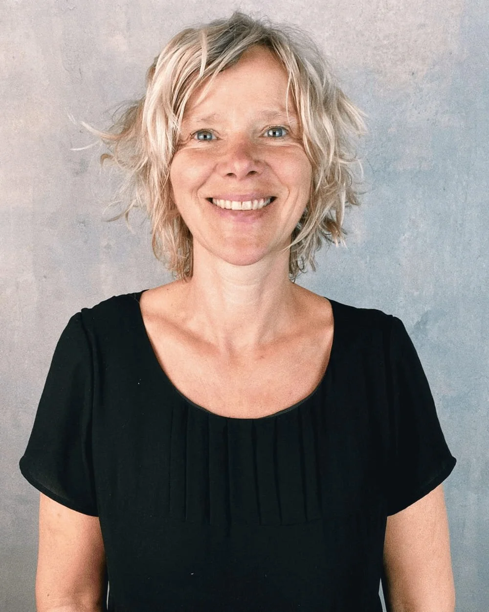 Salon founder Anja with wavy grey-blonde hair, smiling against a textured grey background.