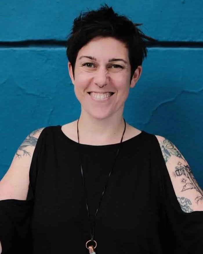Portrait of Julia with short dark hair and tattoos, smiling in front of a blue wall.