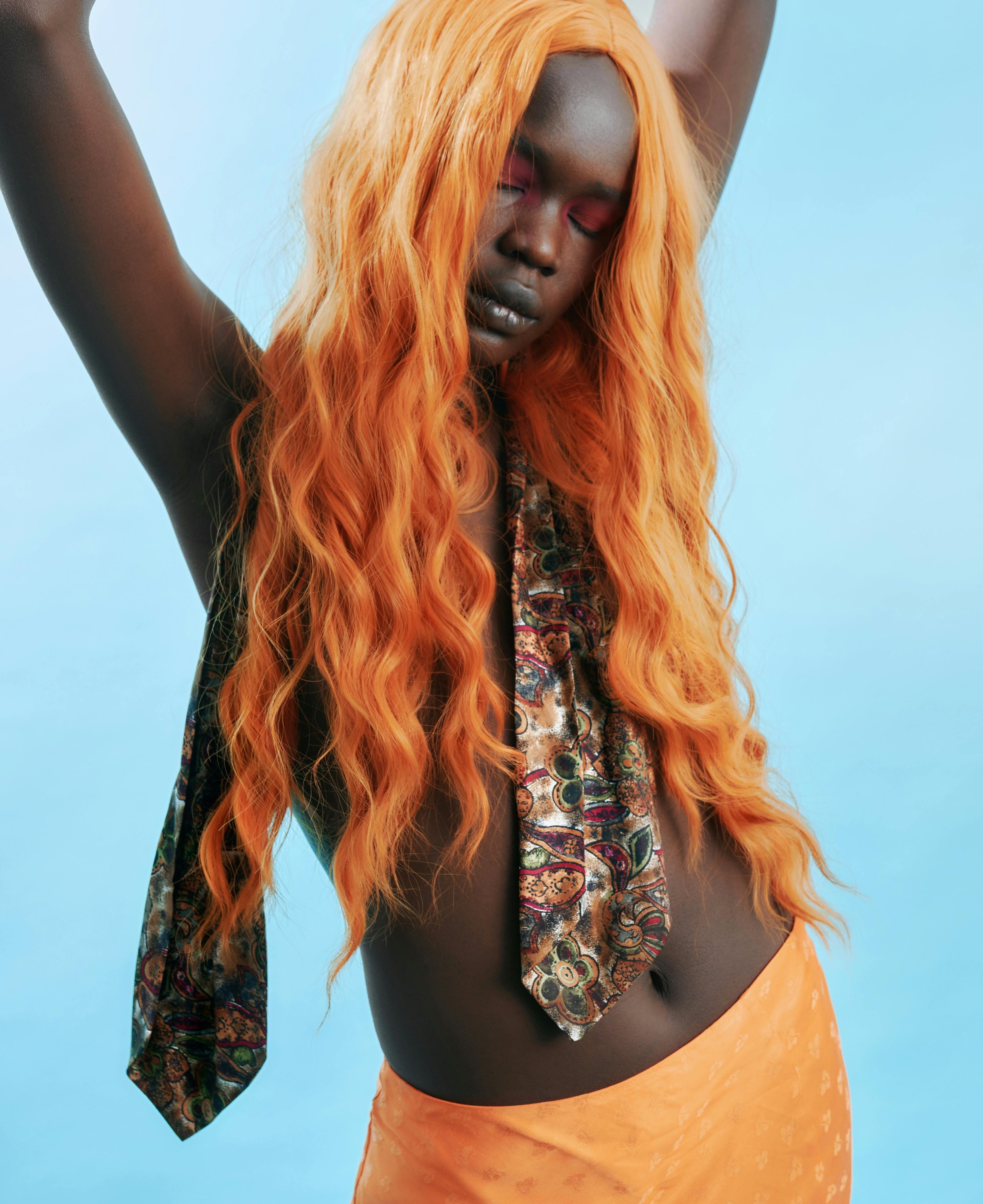 A person of color with long, wavy orange hair, posing with arms raised against a bright blue background.