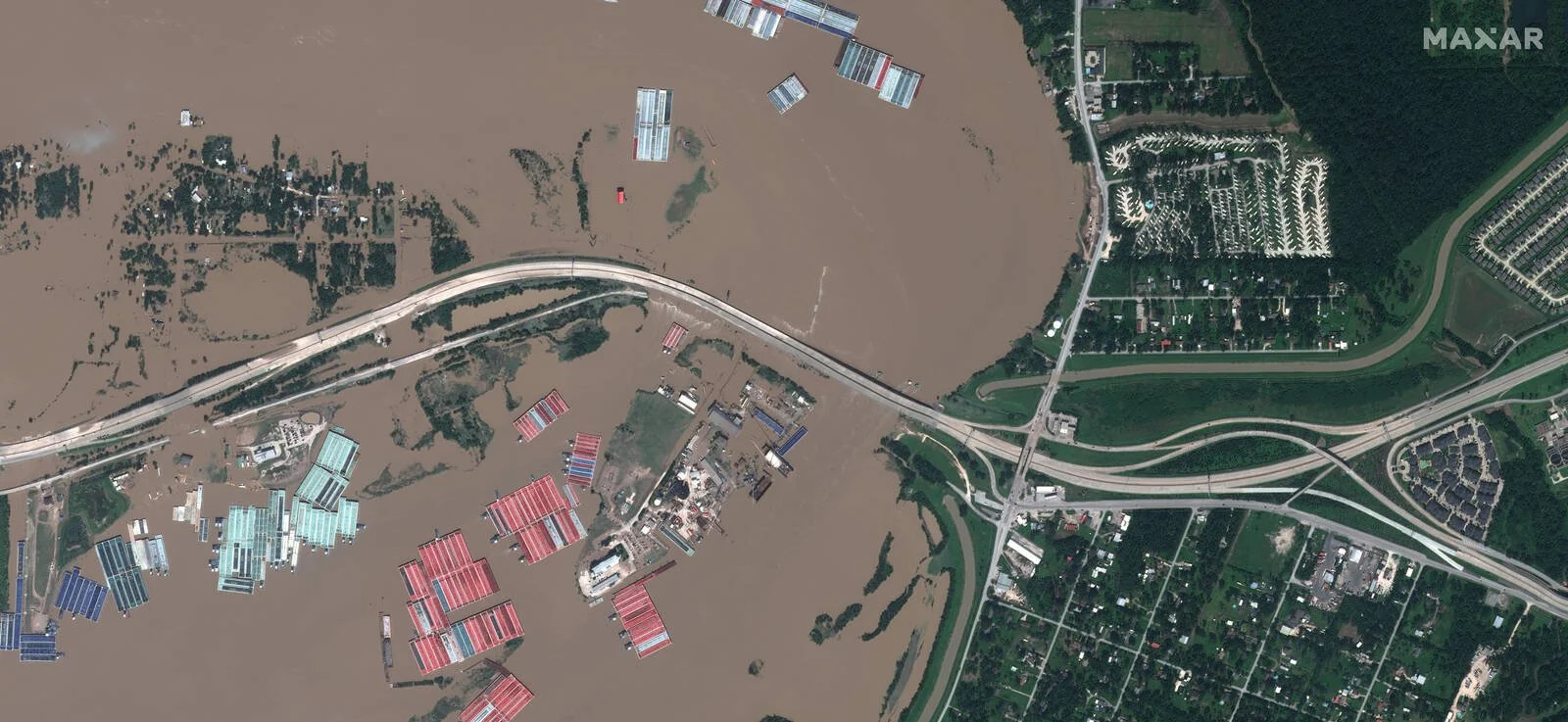 Aerial view showing a flooded area with boats and structures partly submerged, alongside a highway with multiple lanes and curved ramps, and nearby residential neighborhoods.