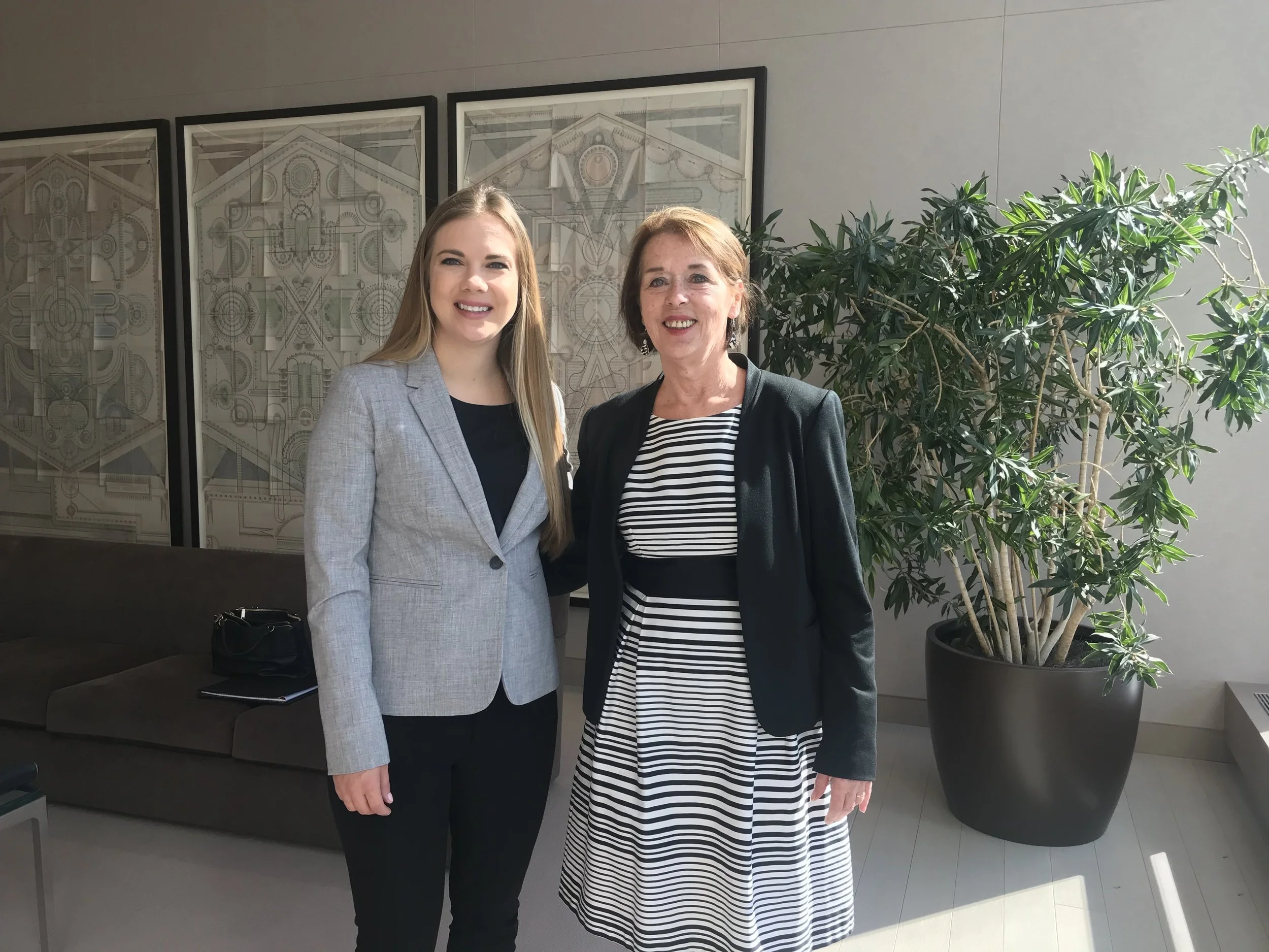 Two women standing together in an office or conference room, smiling at the camera. One woman wears a light gray blazer, the other a black blazer over a striped dress. A large potted plant and framed artwork are visible in the background.