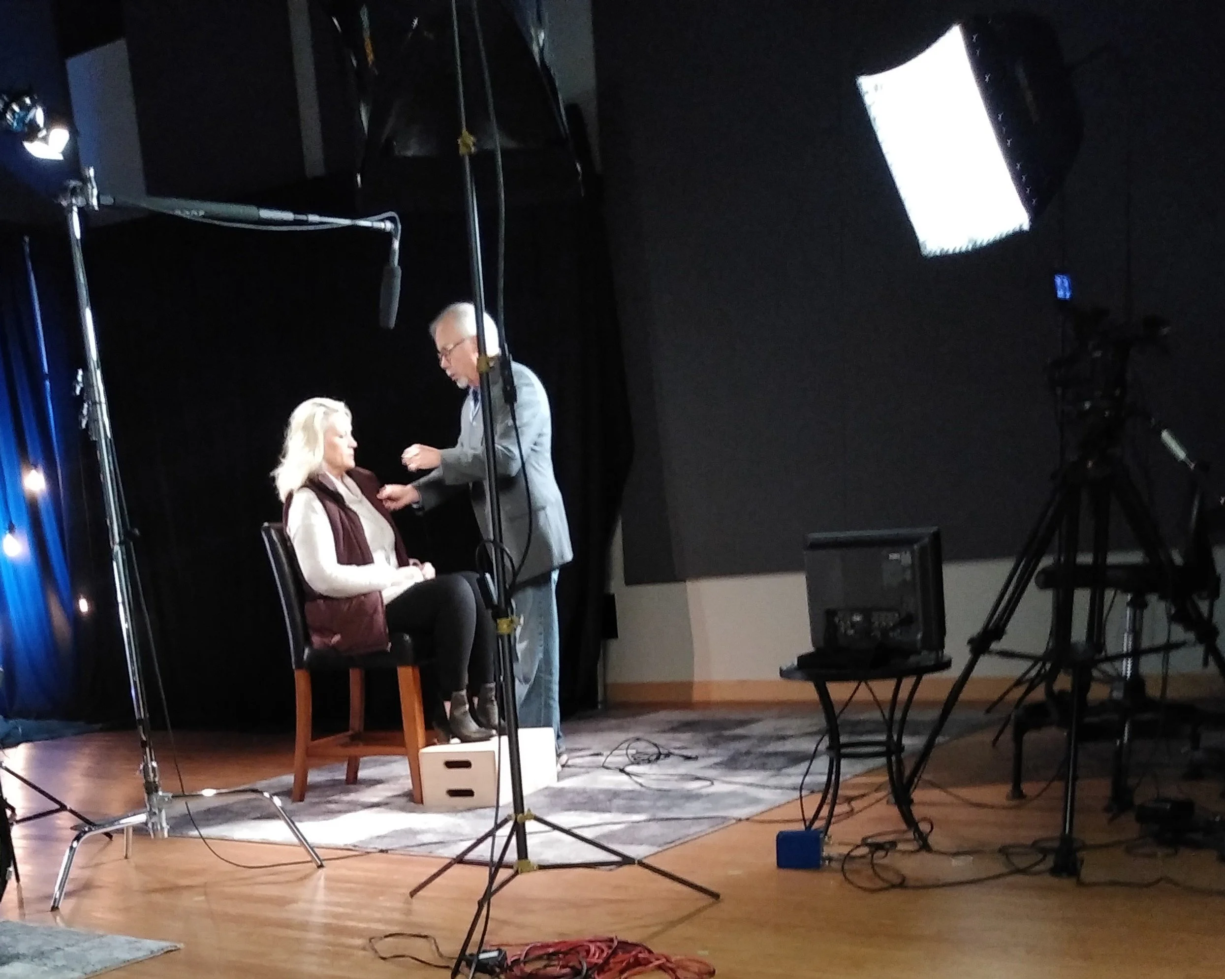 A photo of a recording studio setup with a woman seated on a chair and a man standing next to her, adjusting or pinning something on her shirt. There are studio lights, a camera, and other recording equipment visible.