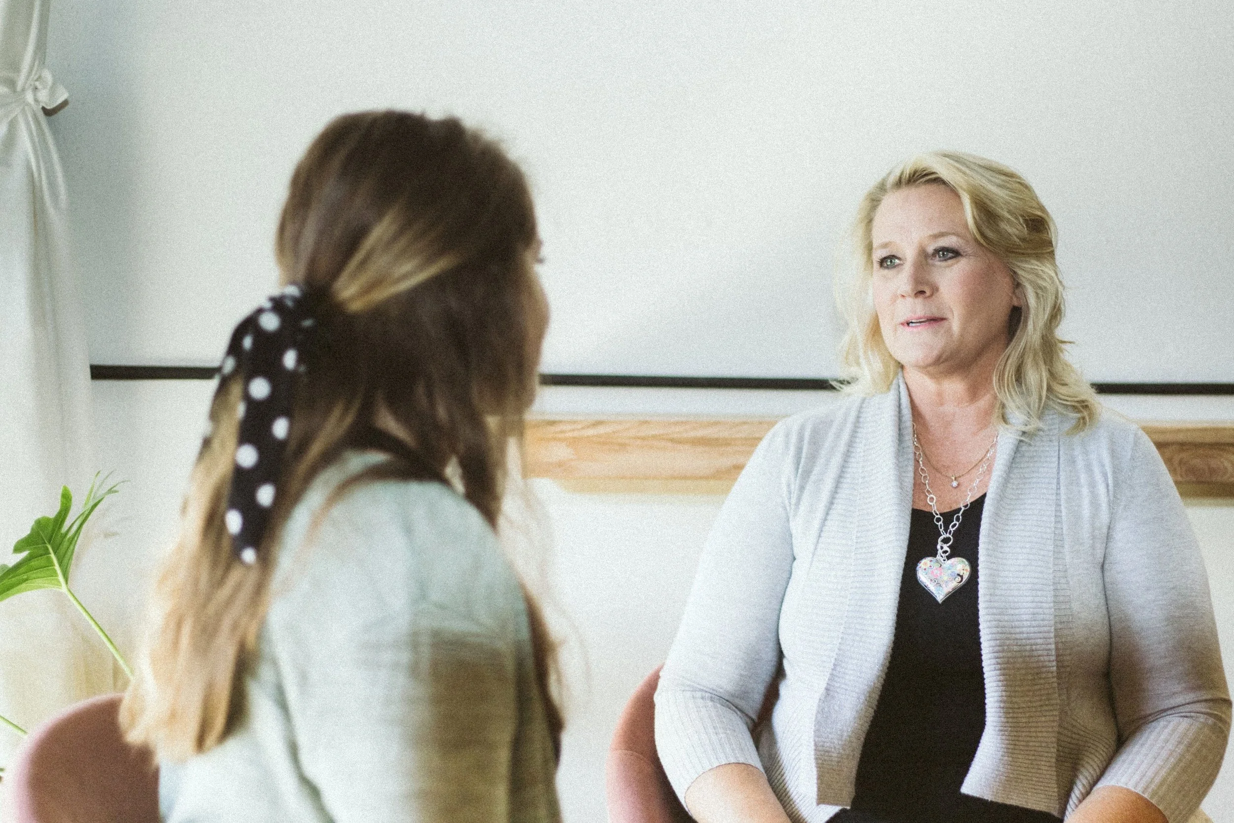Two women having a conversation indoors, one with blonde hair and the other with brunette hair tied with a polka dot scarf, sitting in front of a plain wall and a plant.
