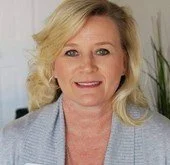 Portrait of a woman with blonde hair wearing a light blue blazer, smiling, standing indoors with plants in the background.