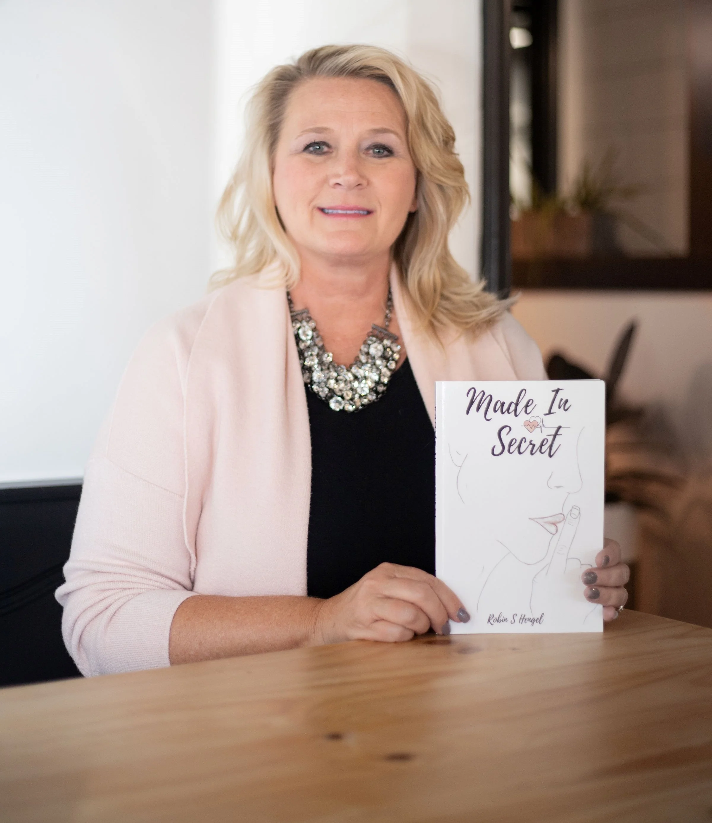 A woman with blonde hair wearing a pink blazer and a statement necklace, sitting at a wooden table, holding a book titled 'Made In Secret' by Robin S. Hooral with a minimalist cover design featuring the outline of a woman's face and finger to lips.