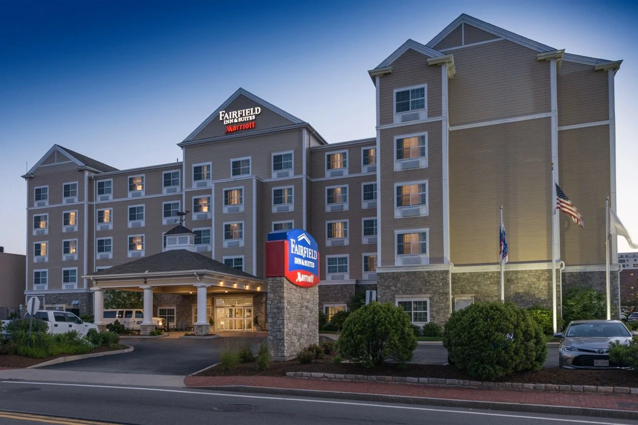 Exterior of a Fairfield Inn & Suites hotel in the evening with a lit sign, flags, trees, and parked cars in front.