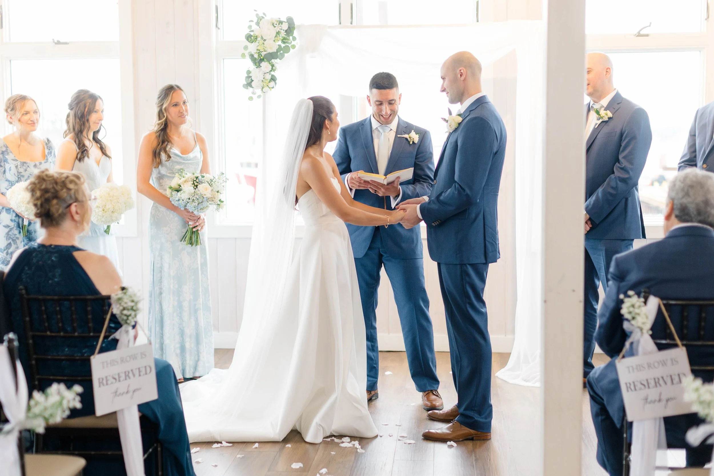 Bride and groom holding hands during wedding ceremony in a bright, decorated room with bridesmaids and groomsmen, with guests seated.
