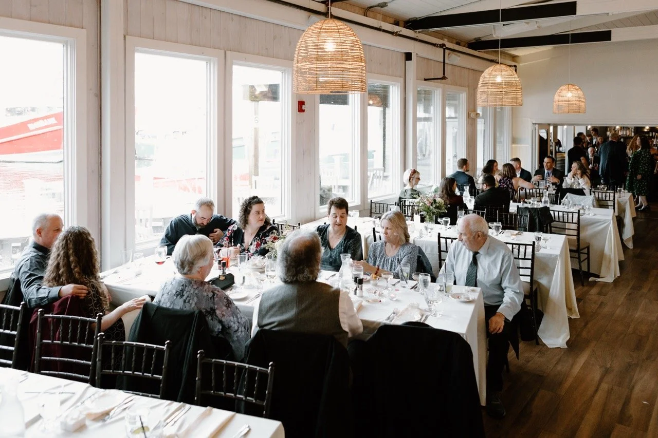 People dining in a well-lit restaurant with large windows, wooden floors, and hanging wicker lamps.