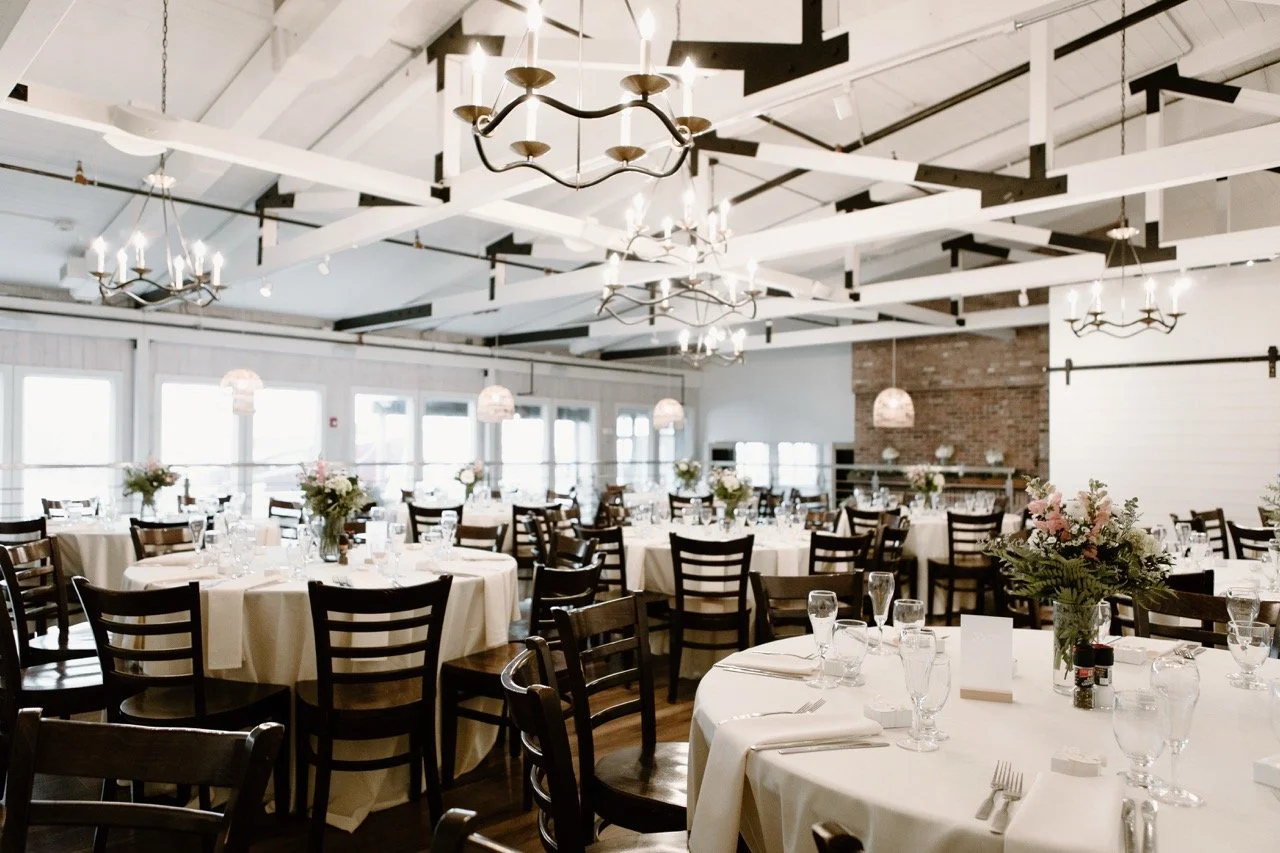 Elegant banquet hall with round tables covered in white tablecloths, set with glassware, utensils, and floral centerpieces, featuring chandeliers and pendant lights hanging from a white ceiling with exposed black beams.