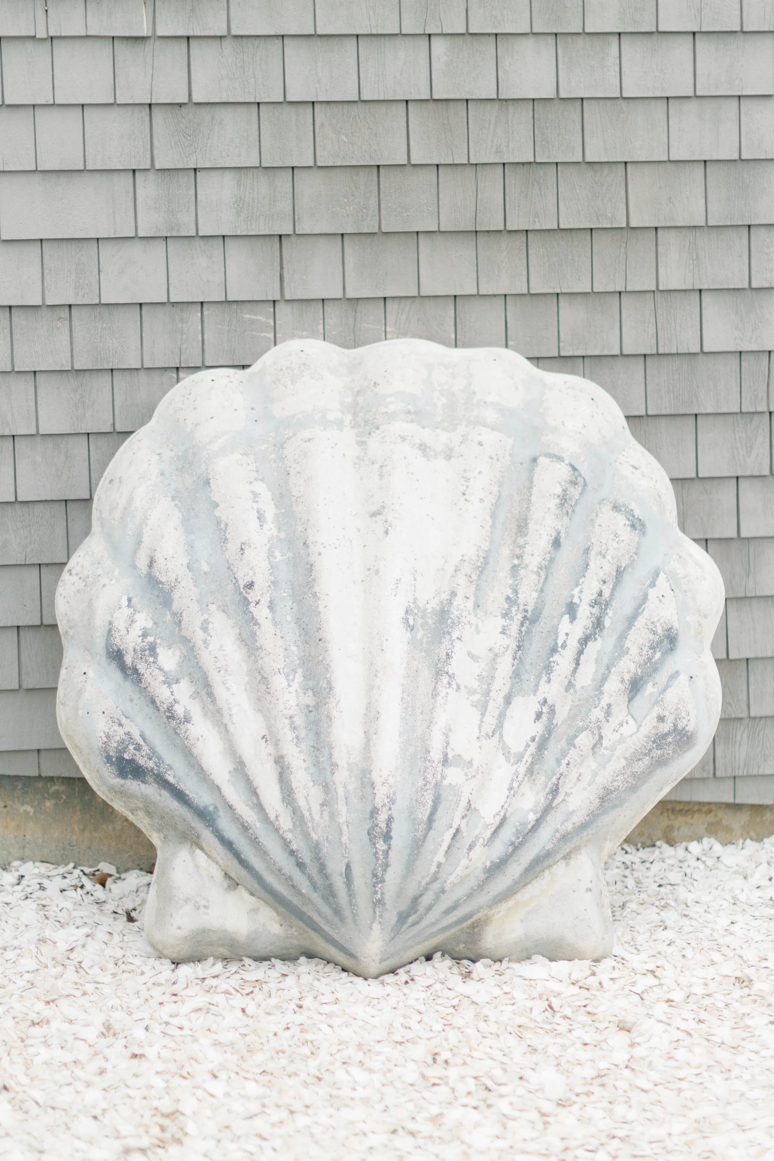 Large decorative shell sculpture in front of a gray shingle wall, situated on a white gravel ground.