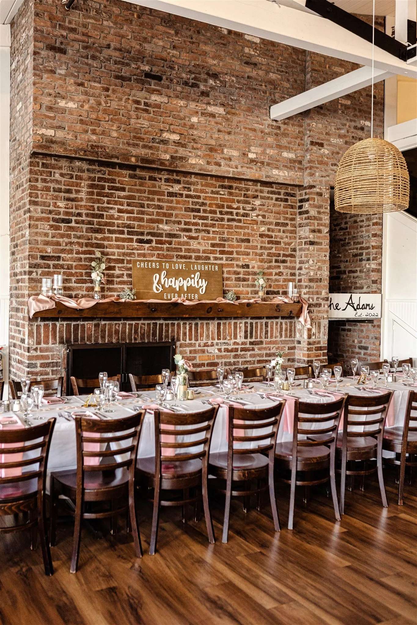A decorated banquet table set for a celebration in a rustic style with a brick wall background, a wooden mantel with a congratulatory sign, and pink ribbons on the table.