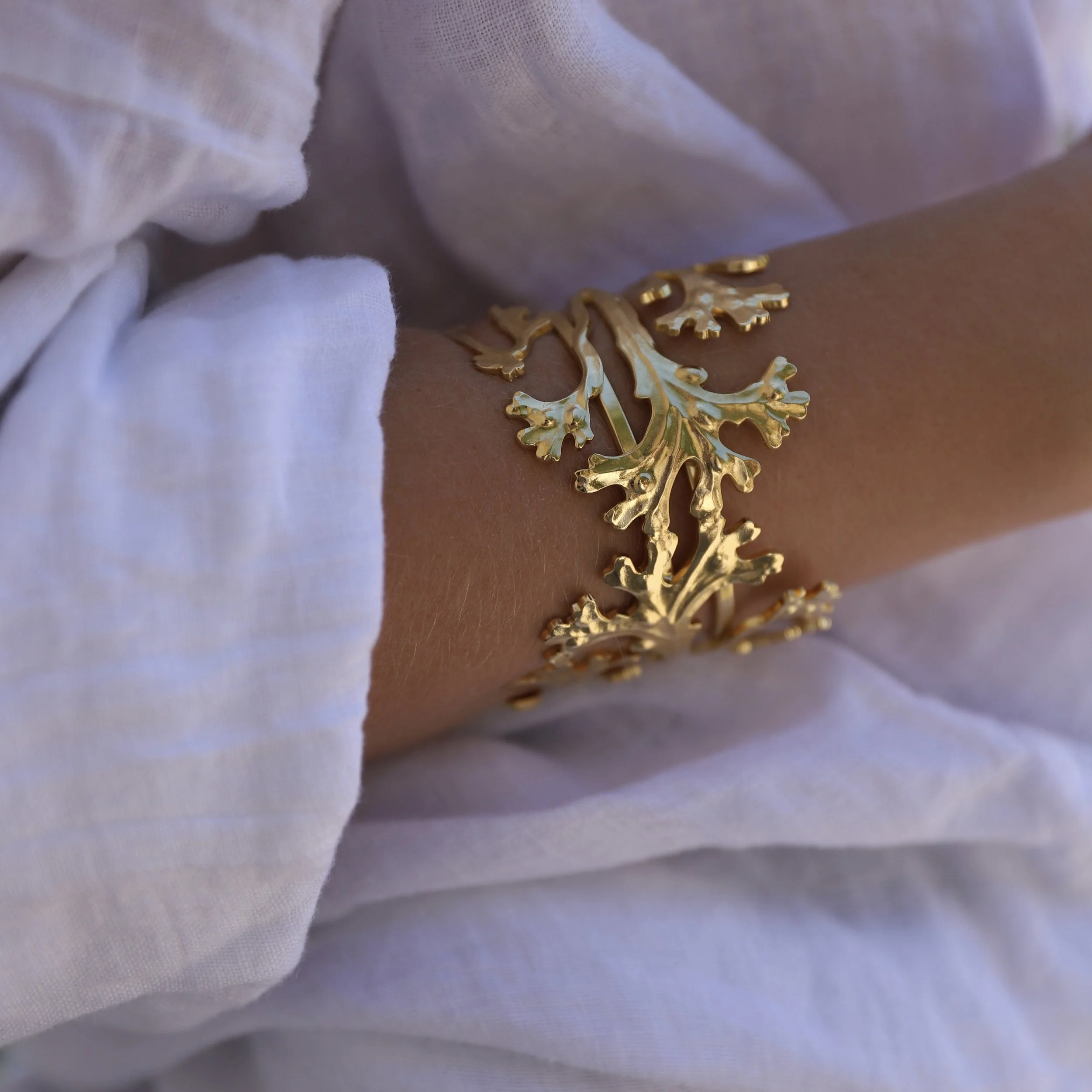 A gold bracelet shaped like tree branches worn on the arm of a person wearing a white coat.