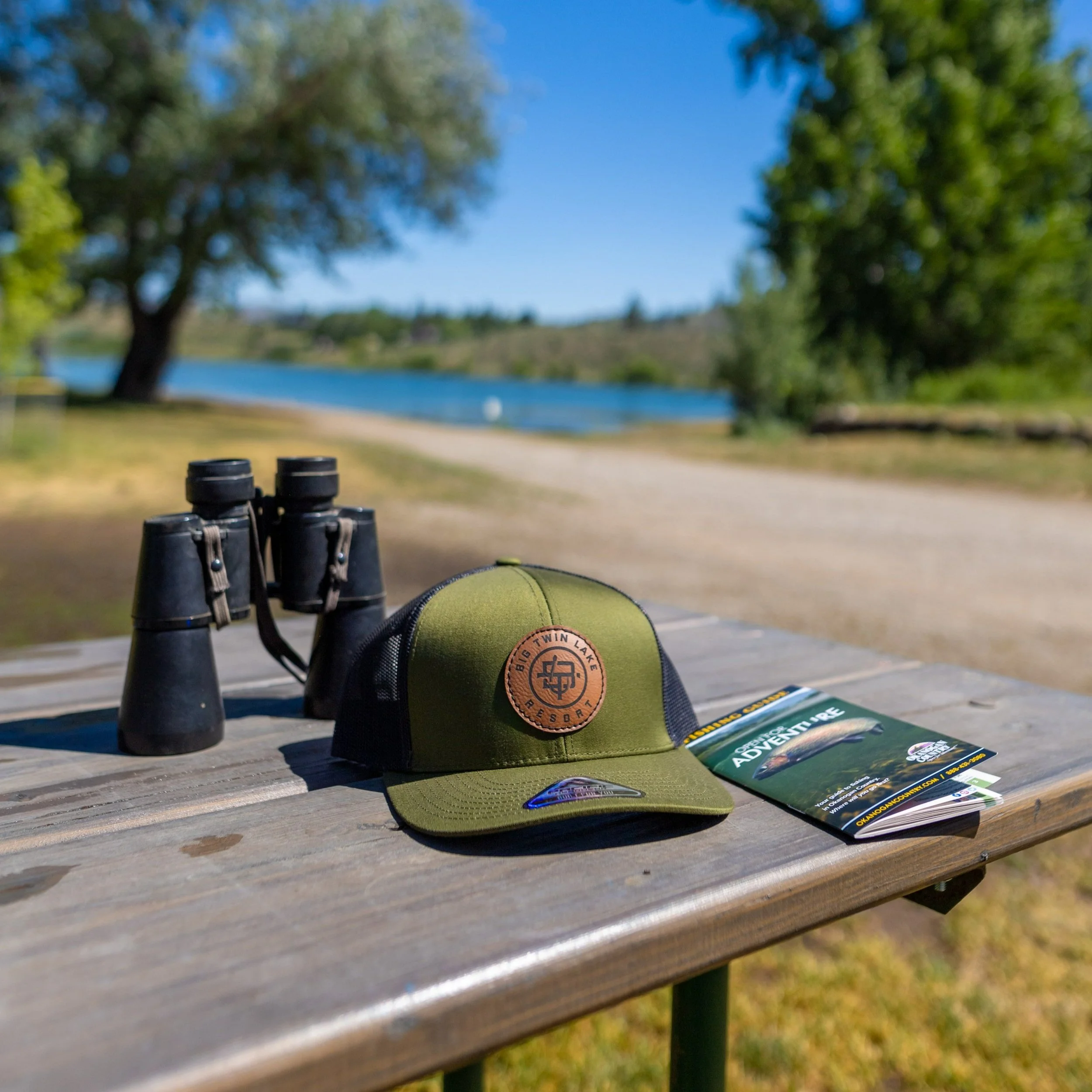 A view of binoculars, a green baseball cap, and camping brochures on a wooden picnic table outdoors near a lake, with trees and a sandy path in the background.