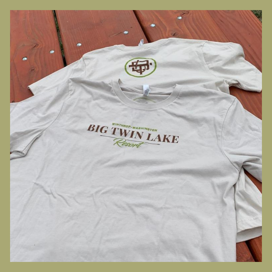 Two white T-shirts laid flat on a wooden surface, one showing text 'Big Twin Lake Resort' and the other with a logo on the back.