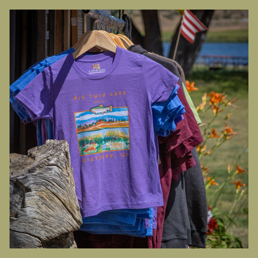 Purple T-shirt with a Big Twin Lake Resort design hanging on a rack outdoors, with other shirts behind it and an American flag in the background.