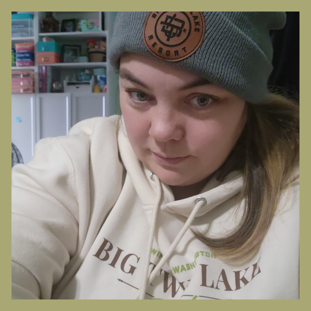 Close-up of a young woman wearing a gray beanie with a leather patch and a cream-colored hoodie with 'Big White Lake' printed on it. She has light-colored eyes and brown hair with blond tips, and is indoors with shelves and storage containers in the