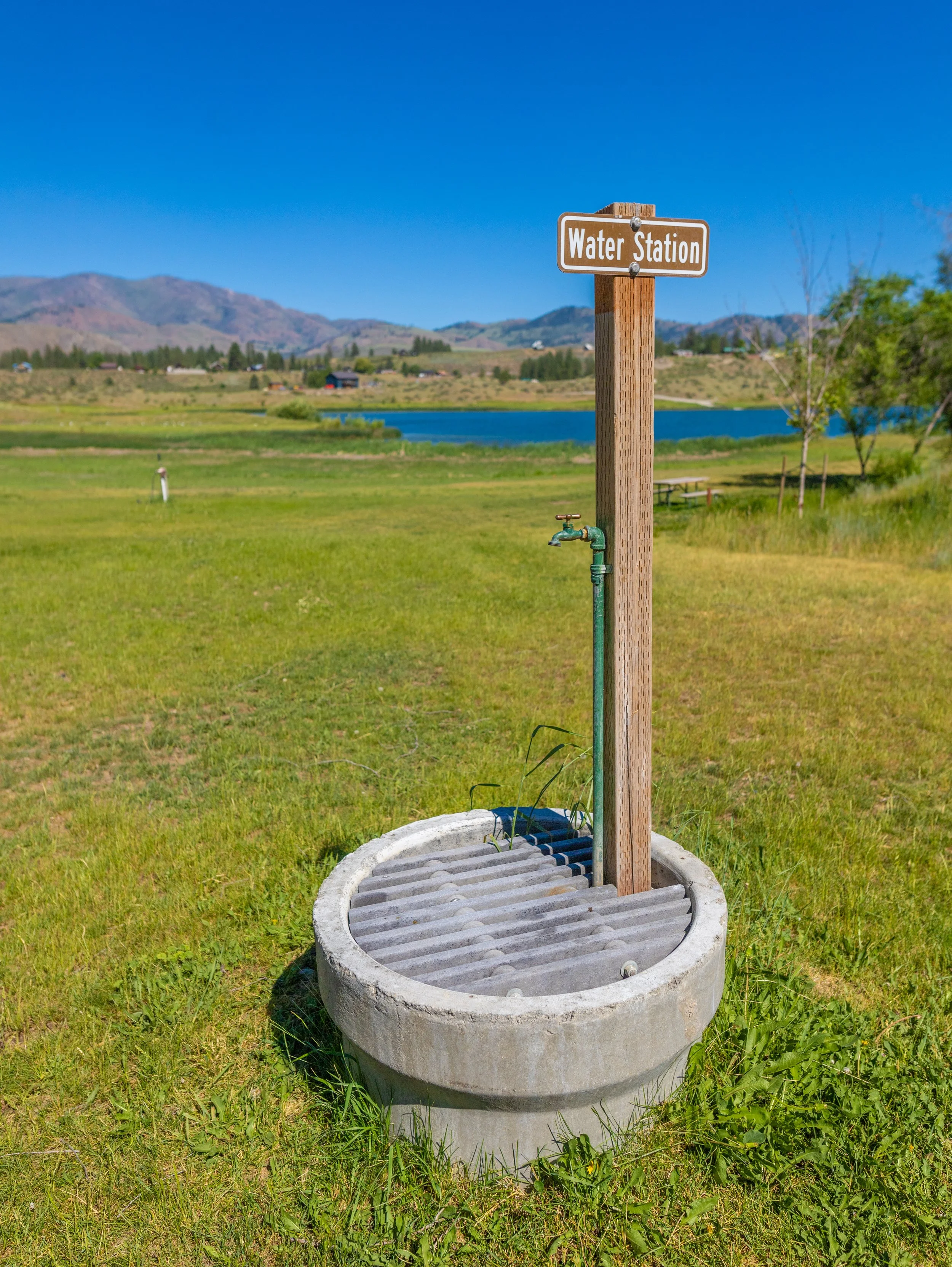 Water fountain with a spigot and a wooden sign that says 'Water Station' set in a grassy area near a lake with mountains in the background.