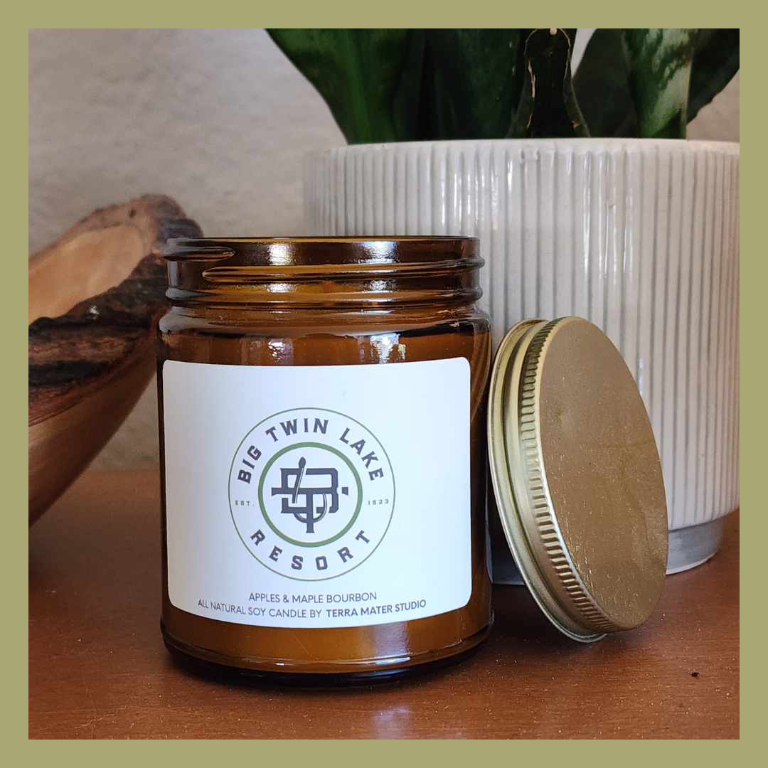 Amber glass jar labeled 'Big Twin Lake Resort' with a gold lid beside a potted plant with striped white planter and a piece of wood in the background.